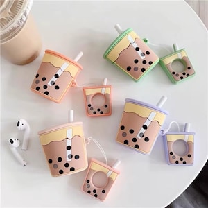 May include: A collection of colorful, bubble tea-themed AirPod cases. The cases are designed to resemble bubble tea cups, with brown, yellow, and pastel-colored accents. Each case has a straw and black boba pearls. White Airpods are also visible.