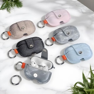 May include: A collection of colorful AirPod cases in various shades, including pink, blue, gray, and patterned fabrics. Each case has a silver keyring and an orange accent. One case is open, revealing white earbuds.