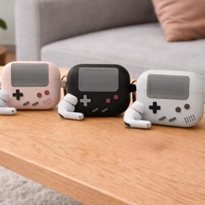 May include: Three AirPod cases in pink, black, and white, each designed to resemble a retro handheld game console. The cases feature a screen, plus and dot buttons, and speaker vents. Each case has a matching pair of white earbuds.