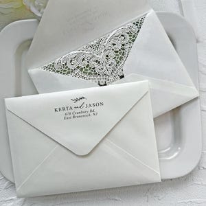 Peut inclure: Enveloppes blanches pour invitations de mariage avec des d&eacute;tails de dentelle complexes et les noms "Kerta and Jason" imprim&eacute;s. Les enveloppes sont dispos&eacute;es sur une assiette blanche, mettant en valeur le design d&eacute;licat et le style &eacute;l&eacute;gant.