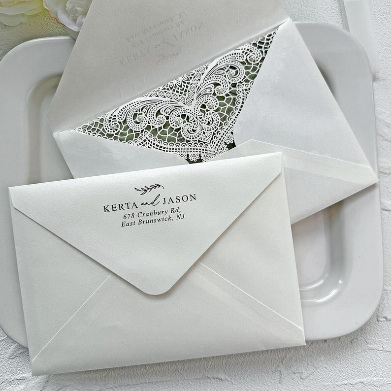 Peut inclure: Enveloppes blanches pour invitations de mariage avec des d&eacute;tails de dentelle complexes et les noms "Kerta and Jason" imprim&eacute;s. Les enveloppes sont dispos&eacute;es sur une assiette blanche, mettant en valeur le design d&eacute;licat et le style &eacute;l&eacute;gant.