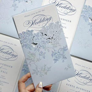 Peut inclure: Faire-part de mariage bleu clair avec un motif floral découpé au laser. L'invitation indique "Please join us for the Wedding of..." en écriture élégante. Le motif présente des roses et des feuilles.