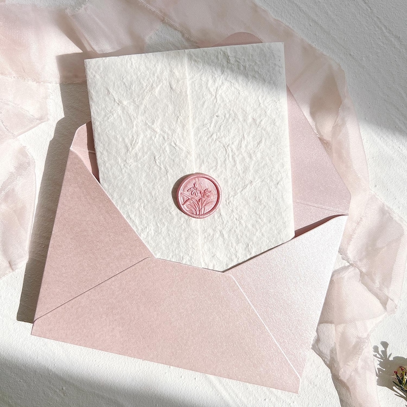Op de afbeelding: Een zachtroze envelop met een getextureerde witte kaart erin, verzegeld met een bijpassend roze waszegel. De zegel heeft een bloemenontwerp. Zachte, transparante stof drapeert rond het briefpapier, wat een delicate en elegante presentatie cre&euml;ert.