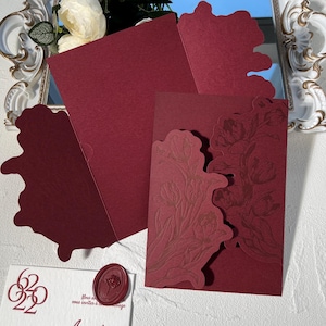 以下が含まれることがあります： バーガンディ色のウェディング招待状セット。セットには、花の模様の招待状、封筒、装飾用のワックスシールが含まれています。招待状は型抜き加工が施されています。セットは白い表面に表示されています。