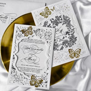 May include: Stylish white wedding invitations with delicate laser-cut floral and butterfly patterns. Gold foil butterflies adorn the cards, which feature the names Amanda and Marc, and the wedding date, August 25, 2023.