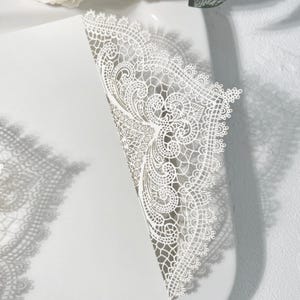 May include: A white, laser-cut invitation with detailed lace-like patterns. The design includes floral and scroll motifs, with a scalloped edge. The invitation is partially open, showing the inside. The shadow of the invitation is visible on the surface.