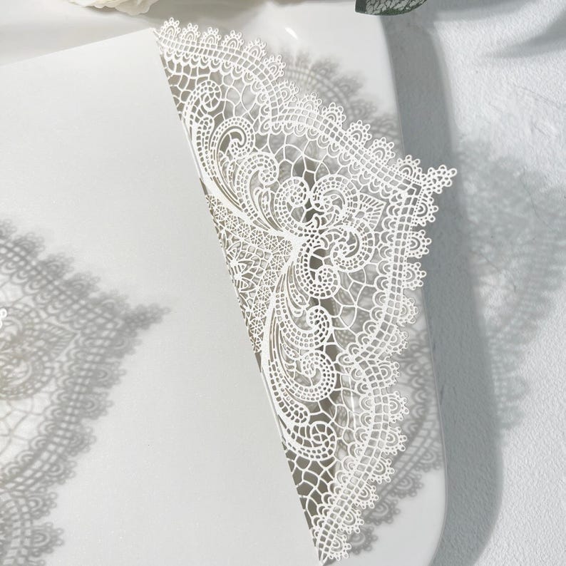 May include: A white, laser-cut invitation with detailed lace-like patterns. The design includes floral and scroll motifs, with a scalloped edge. The invitation is partially open, showing the inside. The shadow of the invitation is visible on the surface.