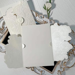 Puede incluir: Juego de invitaciones de boda de color marfil con diseños florales en relieve. El juego incluye una tarjeta con las palabras "THING FANCY", una tarjeta en blanco y elementos decorativos. Un sello de cera y una ramita de vegetación añaden detalles.