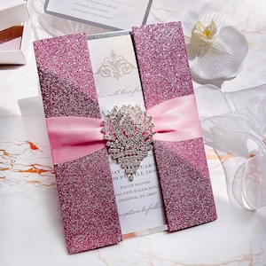 Può includere: Un invito di nozze rosa glitterato con un nastro rosa chiaro e una spilla d'argento. L'invito include dettagli come la data, l'ora e il luogo dell'evento. L'invito è aperto, rivelando i dettagli stampati.