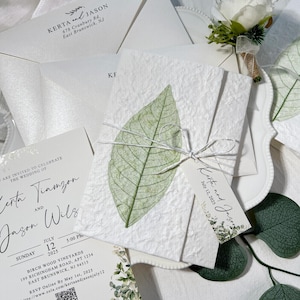 May include: Wedding stationery set with textured white paper, a green leaf, and a tag with names and date. The set includes an invitation, RSVP card, and envelope. The text includes names, date, time, and location.