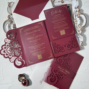 May include: Burgundy wedding invitation suite with gold lettering and decorative laser-cut details. Includes invitation, details card, RSVP card, and envelope. The invitation reads "Rebecca Edwards & William Jackson" and the wedding date.