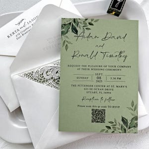 Peut inclure: Une invitation de mariage sur fond vert sauge avec une &eacute;criture &eacute;l&eacute;gante. L'invitation comprend les noms "Aidan David et Ronald Timothy", les d&eacute;tails de l'&eacute;v&eacute;nement et un code QR pour RSVP. Une enveloppe blanche avec un motif en dentelle accompagne l'invitation.