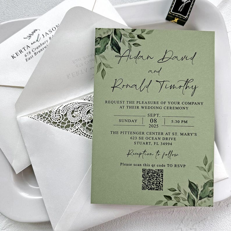Peut inclure: Une invitation de mariage sur fond vert sauge avec une &eacute;criture &eacute;l&eacute;gante. L'invitation comprend les noms "Aidan David et Ronald Timothy", les d&eacute;tails de l'&eacute;v&eacute;nement et un code QR pour RSVP. Une enveloppe blanche avec un motif en dentelle accompagne l'invitation.
