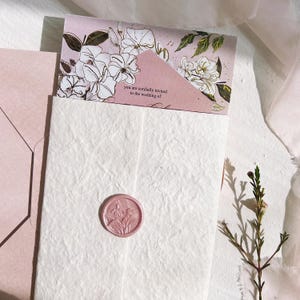 Op de afbeelding: Een trouwuitnodigingsset met een roze envelop en kaart, versierd met bloemenillustraties. De uitnodigingskaart leest "you are cordially invited to the wedding of". Een roze zegel met een bloemenontwerp staat op het witte papier met textuur.