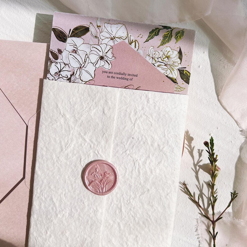 Op de afbeelding: Een trouwuitnodigingsset met een roze envelop en kaart, versierd met bloemenillustraties. De uitnodigingskaart leest "you are cordially invited to the wedding of". Een roze zegel met een bloemenontwerp staat op het witte papier met textuur.