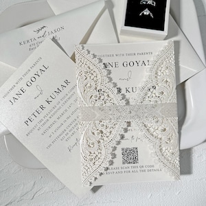 Puede incluir: Elegantes invitaciones de boda blancas con intrincados detalles de encaje. Las invitaciones presentan los nombres de Jane Goyal y Peter Kumar, junto con los detalles del evento. Incluye sobres y un código QR para RSVP.