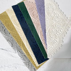 May include: A selection of laser-cut paper in shades of gold, silver, cream, navy, green, lilac, and white. The papers display detailed floral and paisley patterns, suitable for invitations or craft projects. The papers are arranged in a fan shape, showcasing the intricate designs.