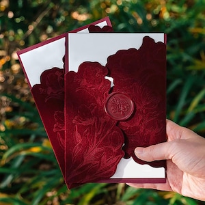 Puede incluir: Invitaciones de boda burdeos con diseño floral y sello de cera. Las invitaciones tienen un borde cortado con láser y un interior blanco. El diseño presenta patrones florales en relieve. Las invitaciones se sostienen en una mano.