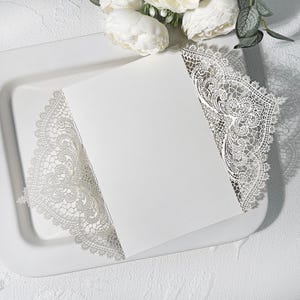 May include: A white invitation card with delicate, lace-style cut-out detailing around the edges. The card rests on a white tray, with white flowers in the background. The design is elegant and suitable for weddings or special occasions.