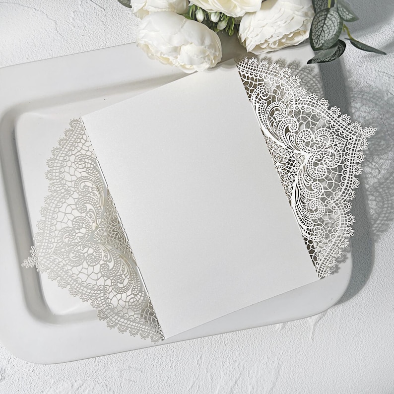 May include: A white invitation card with delicate, lace-style cut-out detailing around the edges. The card rests on a white tray, with white flowers in the background. The design is elegant and suitable for weddings or special occasions.