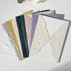 May include: A selection of laser-cut invitation cards in a range of colours, including gold, silver, navy, green, lavender, and cream. The cards display intricate lace-like patterns and are arranged in a fan shape, highlighting their detailed craftsmanship.