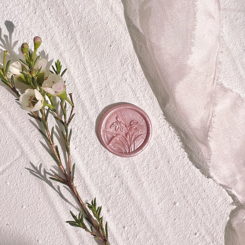 Op de afbeelding: Een roze waszegel met een bloemmotief ligt op een witte, getextureerde ondergrond, naast een takje met witte bloemen en groene bladeren. Een lichtroze stof is op de achtergrond gedrapeerd. De zegel is rond en gedetailleerd.