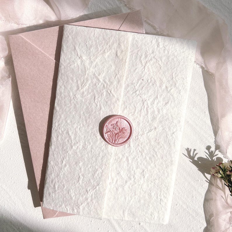 Op de afbeelding: Een briefpapier set met een getextureerde witte papieren kaart, een roze waszegel en een roze envelop. De waszegel heeft een bloemenontwerp. De set ligt op een wit oppervlak met een zachte roze stof op de achtergrond.