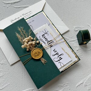 May include: An elegant wedding invitation suite featuring a dark green card with a gold wax seal and dried floral accents. The invitation includes the words "kindly reply" and the initials "KA". A small green ring box is also present.