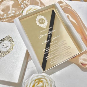 May include: A gold-colored wedding invitation with white text and a black ribbon, presented in a white box. The invitation features ornate design elements and the names Jonny Radeva and Mark Jordan, with the date 17.05.2024.