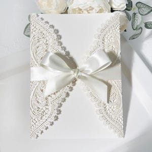 May include: A white invitation card featuring delicate, lace-style cut-out detailing around the borders. A satin ribbon is tied in a bow around the centre. Ideal for weddings or special occasions.
