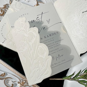 Puede incluir: Elegante juego de invitaciones de boda con un diseño floral en relieve. La tarjeta de invitación tiene las palabras "just love" y los detalles del evento. El juego incluye una superposición decorativa troquelada y un sobre a juego. La paleta de colores es blanco y gris.