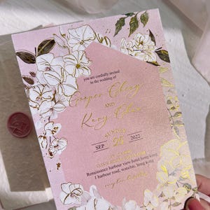 Op de afbeelding: Een roze trouwkaart met gouden folietekst en bloemmotieven. De uitnodiging bevat de namen "Cooper Chezy" en "Kary Colas", en de datum "25 september 2022". De locatie van de bruiloft is ook opgenomen.