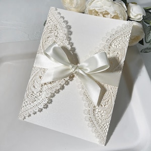May include: Ivory wedding invitation with detailed lace edging. A satin ribbon is tied in a bow around the invitation. The invitation is on a white surface.