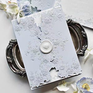 Puede incluir: Una invitación azul claro con diseños florales en relieve y un sello de cera de concha marina. La invitación está colocada sobre una bandeja plateada, con flores blancas y papelería en el fondo. El tema general es elegante y romántico.