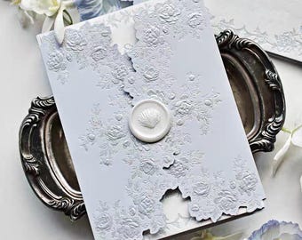 May include: A light blue invitation with embossed floral designs and a seashell wax seal. The invitation is placed on a silver tray, with white flowers and other stationery visible in the background. The overall theme is elegant and romantic.