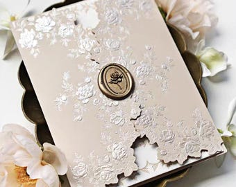 May include: A beige wedding invitation with embossed floral designs and a gold wax seal featuring a rose. The invitation is surrounded by white flowers and rests on a decorative gold tray, creating an elegant presentation.