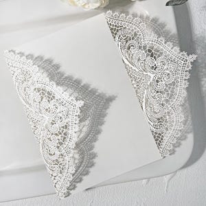 May include: A white invitation card with detailed, lace-style cut-out patterns. The card is open, showing a plain interior. The design includes delicate floral motifs and a scalloped edge, giving a romantic and elegant appearance.