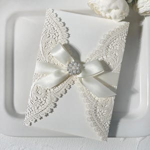 May include: Ivory wedding invitation with detailed lace edging and a satin ribbon bow. The bow features a pearl and rhinestone brooch. The invitation is presented on a white tray.
