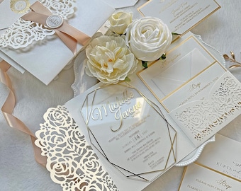 May include: Elegant wedding invitation suite featuring white and gold accents. Includes laser-cut envelopes, RSVP cards, and a clear acrylic invitation with gold lettering. Decorative floral elements and ribbon add to the sophisticated design.