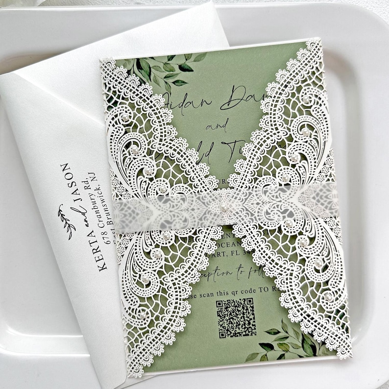 Peut inclure: Faire-part de mariage avec un motif de dentelle blanche d&eacute;coup&eacute;e au laser sur un fond vert. L'invitation comprend les noms "Aidan Dan and ..." et un code QR. Une enveloppe blanche avec les noms "Kerta and Jason" est &eacute;galement visible.