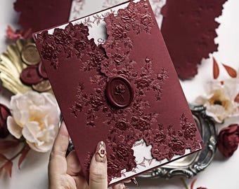 Könnte beinhalten: Bordeauxfarbene Hochzeitseinladung mit floralen Ausschnitten und einem Wachssiegel. Die Einladung weist geprägte Blumendesigns und einen dekorativen Rand auf. Die Einladung wird in einer Hand gehalten, mit anderen Einladungen und floralen Dekorationen im Hintergrund.