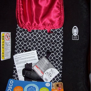 May include: A black and white patterned car seat with a red satin insert. The seat is labeled "Cosco Scenara Next Convertible Car Seat" and features side impact protection. The seat is designed to fit three children across the back seat. The seat pad is washable.