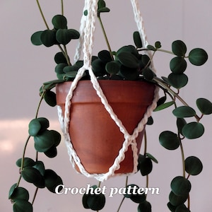 May include: A terracotta-colored plant pot suspended by a white macrame plant hanger. The pot holds a green leafy plant with round leaves. The words "Crochet pattern" are visible at the bottom of the image. The plant's vines cascade down.