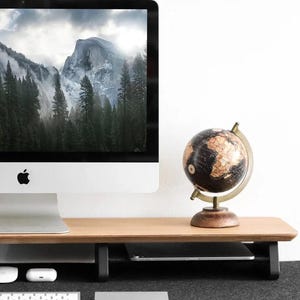 May include: A wooden monitor stand with a black metal frame and a globe on top. The stand has a shelf underneath for storing items like a keyboard or tablet. The stand is on a desk with a black mat.