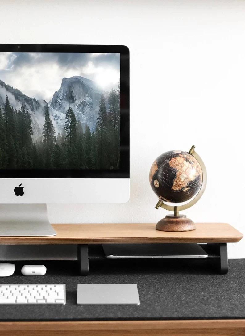 May include: A wooden monitor stand with a black metal frame and a globe on top. The stand has a shelf underneath for storing items like a keyboard or tablet. The stand is on a desk with a black mat.