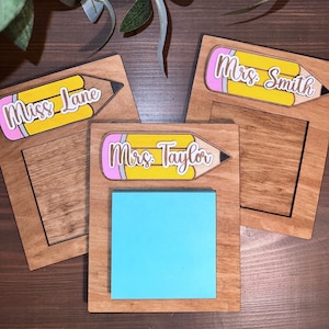 May include: Three wooden note holders with a pencil design and a space for sticky notes. The pencils are yellow with pink erasers and have the names "Miss Lane", "Mrs. Taylor", and "Mrs. Smith" written on them.