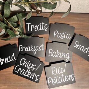 May include: Black and white labels with handwritten text for organizing kitchen pantry items. The labels include "Treats", "Pasta", "Snacks", "Breakfast", "Baking", "Bread", "Chips/Crackers", and "Rice/Potatoes".