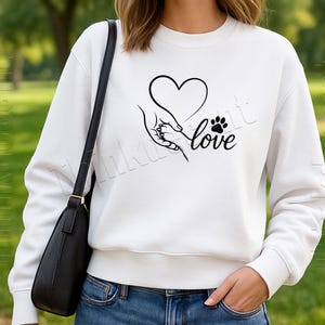 May include: White sweatshirt with a black graphic of a heart, a hand holding a paw, and the word "love". The sweatshirt is a crew neck style. The person is wearing blue jeans and a black purse.