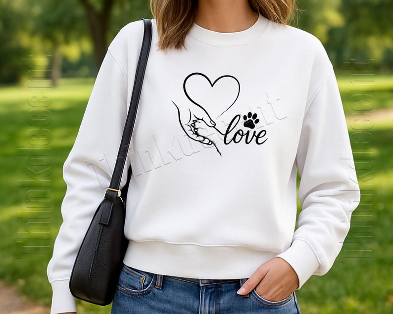 May include: White sweatshirt with a black graphic of a heart, a hand holding a paw, and the word "love". The sweatshirt is a crew neck style. The person is wearing blue jeans and a black purse.