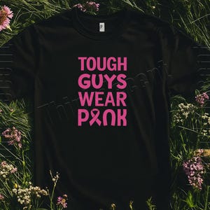 Puede incluir: Camiseta negra con el texto "TOUGH GUYS WEAR PINK" en letras rosas brillantes. El diseño incluye un símbolo de cinta rosa. La camiseta se muestra sobre un lecho de hierba verde y flores.
